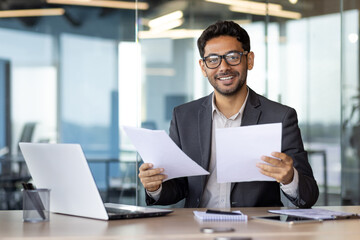 Portrait of successful arab financier, businessman in glasses smiling and looking at camera, man inside office behind paperwork holding documents and financial reports, satisfied with achievements.