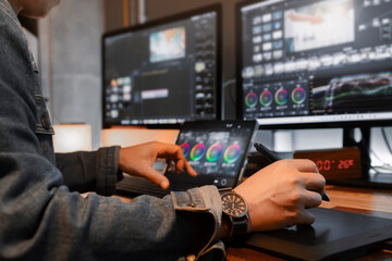 close-up of an Asian male movie video editor or colorist, wearing denim jacket, working with footage or video on his personal computer by pen tool and tablet, in his creative office studio. - obrazy, fototapety, plakaty