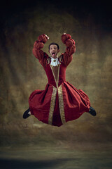 Young emotional man, medieval royal person emotionally jumping in female dress against vintage background. Concept of historical retrospectives, fashion, provoking projects, gender fluidity