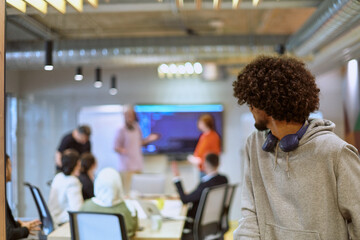 Fototapeta premium In a modern office environment, an African American young entrepreneur with headphones engages in work, while in the background, his dedicated colleagues exemplify teamwork and collaboration