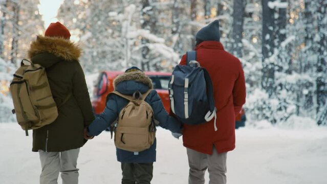 Back view of parents and son in warm clothes approaching car while having road trip in winter