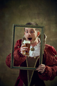 Medieval royal person, prince in female dress holding picture frame and drinking beer against dark green background. Concept of historical retrospectives, fashion, provoking projects, festival