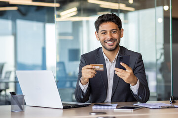 Portrait of young successful Asian businessman inside office, man looking at camera smiling, using online application on phone for internet shopping, holding bank credit card in hand.