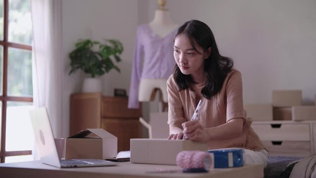 young Asian woman SME startup small business freelance writing a customer's shipping address on parcel box before sending it to customer, SME e-commerce concept.