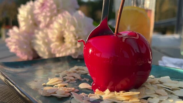 in a restaurant on the table there is a beautiful dessert in the form of a large cherry berry with a spoon breaking a mousse dessert on almond flakes. in the background a glass with an orange drink