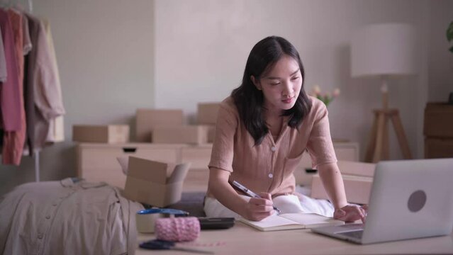 Online Asian Women entrepreneur merchant concept, Fashion designer wearing casual clothing checks online cloth shopping order and working on laptop