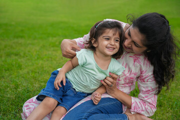 Happy playful young indian mother and cute little girl daughter sitting at summer park or garden having fun together.