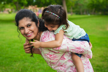 Happy young indian child daughter playing piggy back ride with her mother at summer park Family and...