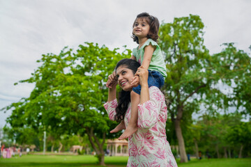 Happy young indian mother carrying child daughter on her shoulders at summer park.Family and...