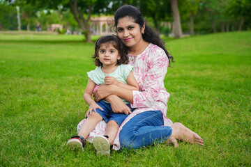 Naklejka na ściany i meble Happy young indian mother and cute little girl daughter sitting at summer park or garden.