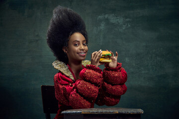 Beautiful african woman, medieval princess eating delicious burger against vintage green background. Fast food. Concept of history, beauty and fashion, comparison of eras, ad