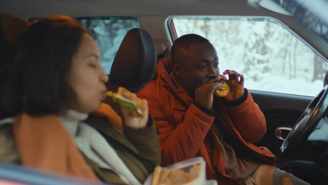 Selective focus on black man eating sandwich with his wife sitting on passengers seat next to him in warm clothes during winter trip