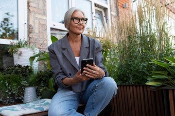 Obraz premium stylish mature woman with gray hair and glasses dressed in a gray jacket chatting in a smartphone on the street