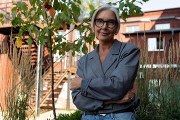 a slender European gray-haired senior business woman with a stylish hairstyle and glasses is dressed in a gray jacket against the backdrop of urban architecture