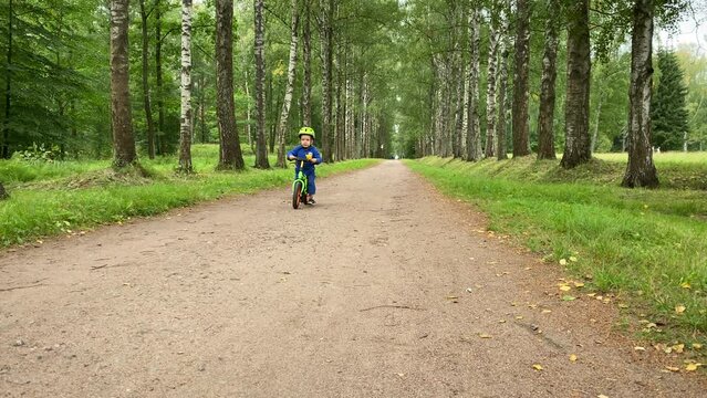 toddler child in safety helmet riding balance bike in summer forest, little boy rides bike in the park