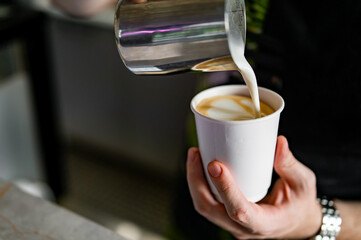 Professional barista hands pouring steamed milk into coffee paper cup making coffee.