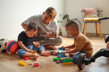 Cheerful young father playing ukulele sons playing with toys at home - obrazy, fototapety, plakaty