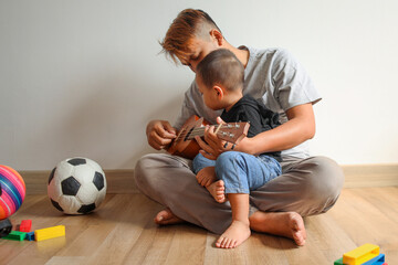 Father sitting cross legged on floor and playing ukulele with his son at home