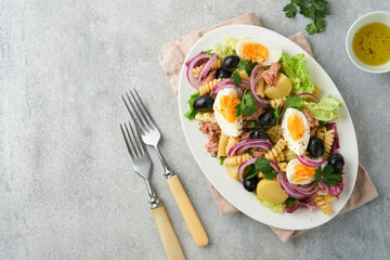 Tuna salad with pasta, eggs, potatoes, olives, red onions and sauce in white plate on old light gray concrete table background. Nicoise salad. French cuisine. Top view. Flat lay.