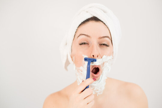 Cheerful caucasian woman with a towel on her head and shaving foam on her face holds a razor on a white background