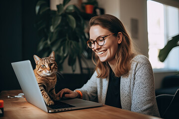 Carefree freelancer girl with glasses smiles while working at a laptop with a cat at the workplace - obrazy, fototapety, plakaty