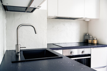 Kitchen counter top, faucet, water tap and sink. White and black Scandinavian interior design in modern apartment and home. Induction cooker stove and electric oven in the background.