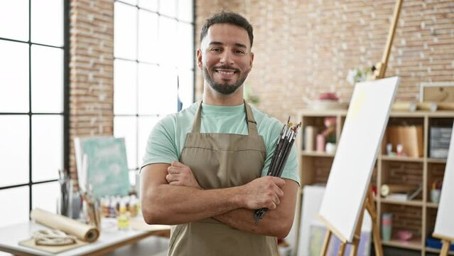 Young arab man artist standing with arms crossed gesture holding paintbrushes smiling at art studio