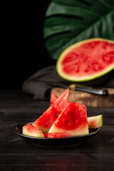 Close-up of watermelon pieces on wooden table with open watermelon and green leaf in the background out of focus, black background, vertical