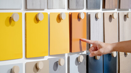 Designer hand pointing at orange laminate sheet on material swatch wall display in material library. Choosing color board from collection for interior architecture and furniture finishing design - obrazy, fototapety, plakaty