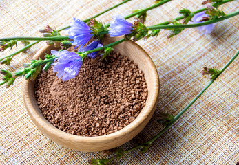 Ground chicory root in a wooden cup and chicory flowers on a rustic wooden background. Alternative...