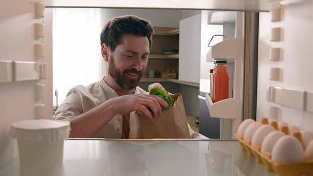 Point of view POV from inside refrigerator middle-aged adult man guy happy smiling at home kitchen taking goods from paper bag put products in fridge eggs lettuce milk pepper shopping food delivery