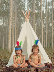 Twins sitting cross-legged in front of a tepee playing at being Native Americans. . © OR MIXphoto
