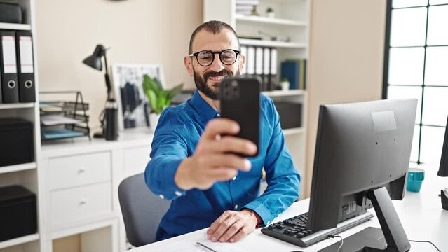 Young hispanic man business worker make selfie by smartphone smiling at office