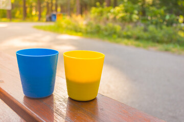 Blue and yellow cups with coffee in morning sunshine. Patriotic ukrainian colours. Two plastic mugs with cappuccino. Portable camping cups. Outdoor dating.  - obrazy, fototapety, plakaty