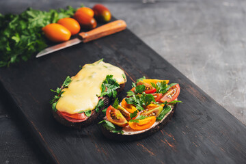 Two sandwiches with toasted bread, arugula, parsley and tomatoes and melted cheese and bacon on a dark wooden board