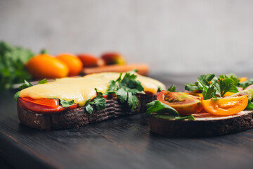 Two sandwiches with toasted bread, arugula, parsley and tomatoes and melted cheese and bacon on a dark wooden board