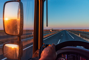 View from the driver's seat of a truck, with a straight road ahead and the sun rising in the rearview mirror.