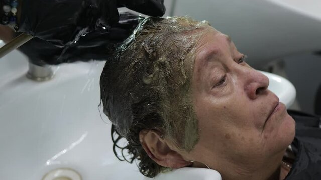 Beginning to remove brown tint from an elderly woman with water on salon washing bed, profile view, grandmother with short hair. Old age fashion and beauty concept