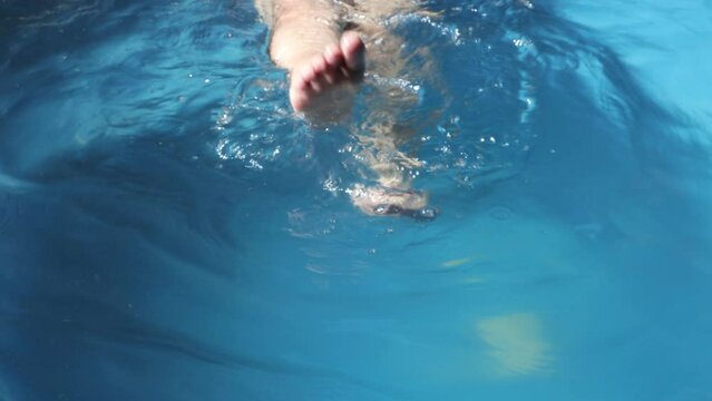 Feet of an adult woman kicking water, splashing and moving relaxed in a swimming pool with blue bottom, reflected sunlight, sunny day in a day spa