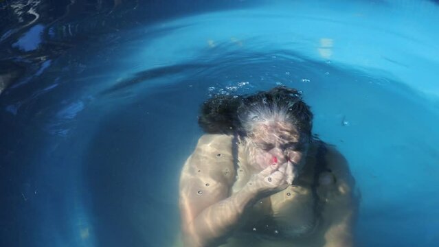 Senior adult Mexican woman in small pool, submerging with hand over nose, outdoor pool, arms moving slightly, blue in background, long wet gray hair, red painted nails, sunny day in Mexico