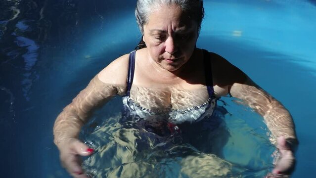 Senior adult Mexican woman relaxing in a small pool, moving her hands in water and looking down, outdoor pool, blue in background, long wet gray hair, red painted nails, sunny day in Mexico