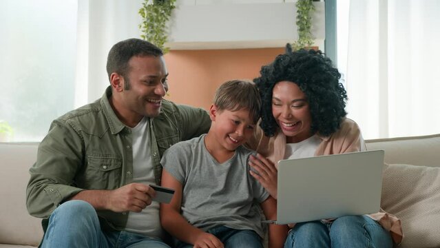 Happy family little boy teen child kid with parents mom and dad on couch shopping online with bank credit card on laptop African American mother and father teach son buying home goods internet payment
