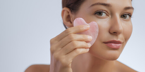 Beauty and skincare concept with a beautiful young adult woman doing a skincare routine, self-massage with the gua sha tool. Female model using a pink gemstone massager for facial lifting. Studio shot © Buyanskyy Production