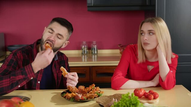 Young hungry brunette man in the kitchen at the table eating chicken wings. A young blonde woman looks bored at it. Eating fast food.