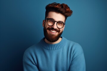 Fototapeta premium Young handsome man with a beard in a sweater and glasses on a blue background, happy face smiling, looking at the camera. A positive person. Generative AI