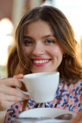 A happy and beautiful young woman enjoying a cup of coffee at a cafe, radiating cheerful and leisurely vibes in a city setting.