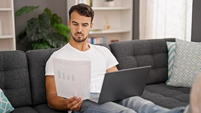 Young hispanic man using laptop reading paper at home