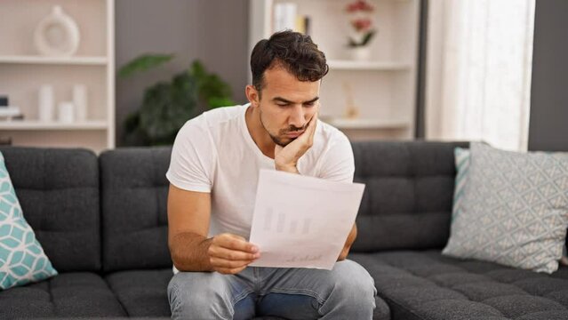 Young hispanic man reading bill sitting on sofa looking upset at home