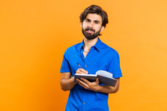 Thoughtful Journalist Young Middle Eastern Man Making Notes, Writing Down Thoughts With Pen Into Notepad Notebook Diary, To Do List, Good Idea Inspiration. Arabian Guy Isolated On Orange Background