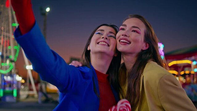 Smiling girls making selfie at illuminated funfair closeup. Two friends hugging